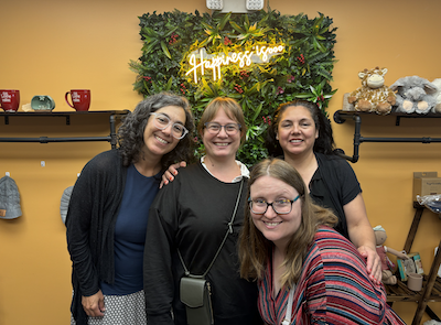 Four women smile at the camera for a posed picture in front of a selfie background made of green leaves and a neon sign that reads "Happiness is..." 